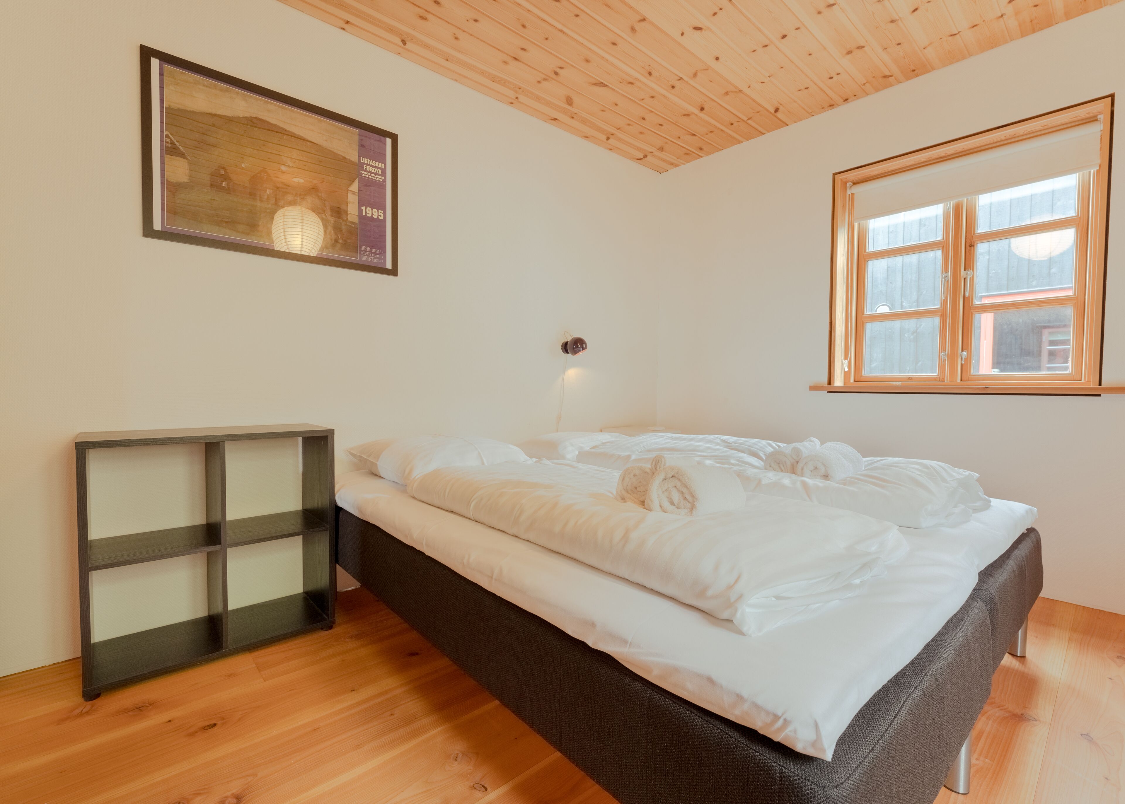 Un dormitorio con techo de madera, cama con ropa de cama blanca, mesita de noche y un cuadro enmarcado en la pared.