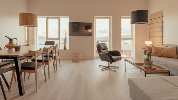 Una sala moderna con comedor, un sofá, televisor montado en la pared y ventanas amplias con vistas.