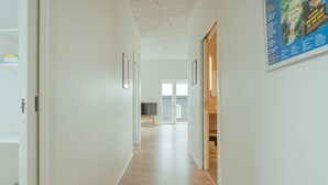 Un couloir doté d’un plancher en bois, de murs blancs et d’une porte donnant sur une pièce où se trouvent un lit et une fenêtre.