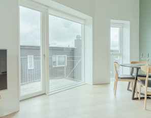 Um apartamento moderno com porta de vidro deslizante que dá para uma varanda, área de jantar com mesa e cadeiras, e vista para a paisagem urbana.