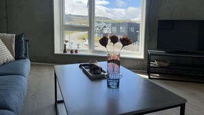 Een woonkamer met een groot raam, een flatscreen-televisie, een bijzettafeltje met een vaas en een dienblad, en een bank met kussens.