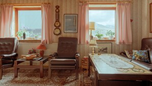 A cozy living room with a brown leather sofa, a brown leather armchair, a wooden coffee table, and a patterned rug. There are two windows with pink curtains, a wall clock, and a framed picture. - Charming 3 Br Home | Scenic Village | Nature (Eiði)