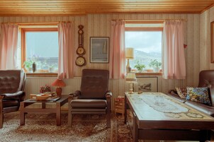 Ein gemütliches Wohnzimmer mit einem braunen Ledersofa, einem braunen Ledersessel, einem Holztisch und einem gemusterten Teppich. Es gibt zwei Fenster mit rosa Vorhängen, eine Wanduhr und ein gerahmtes Bild.