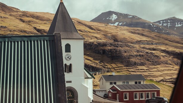 A church with a clock tower, a red house, and a mountainous backdrop. - Charming 3 Br Home | Scenic Village | Nature (Eiði)