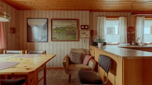 A cozy cabin interior with a wooden dining table, two chairs, a kitchen area with a sink, and a window with curtains. - Charming 3 Br Home | Scenic Village | Nature (Eiði)