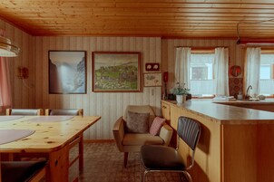 Ein gemütliches Blockhaushaus-Interieur mit einem Holzespeisetisch, zwei Stühlen, einer Küchenzeile mit Spüle und einem Fenster mit Vorhängen.