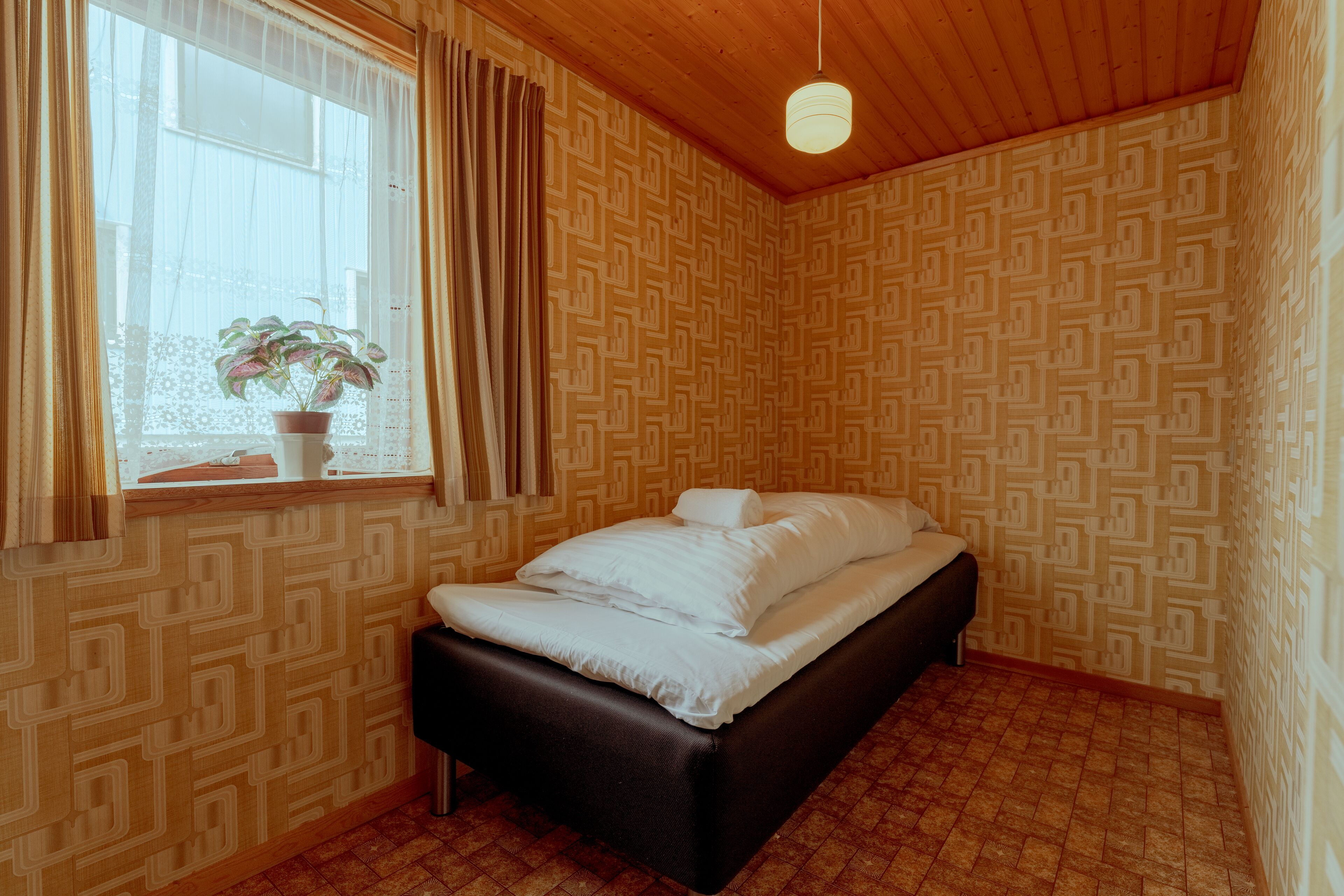 A room with a bed, a window with curtains, a potted plant, and patterned wallpaper.