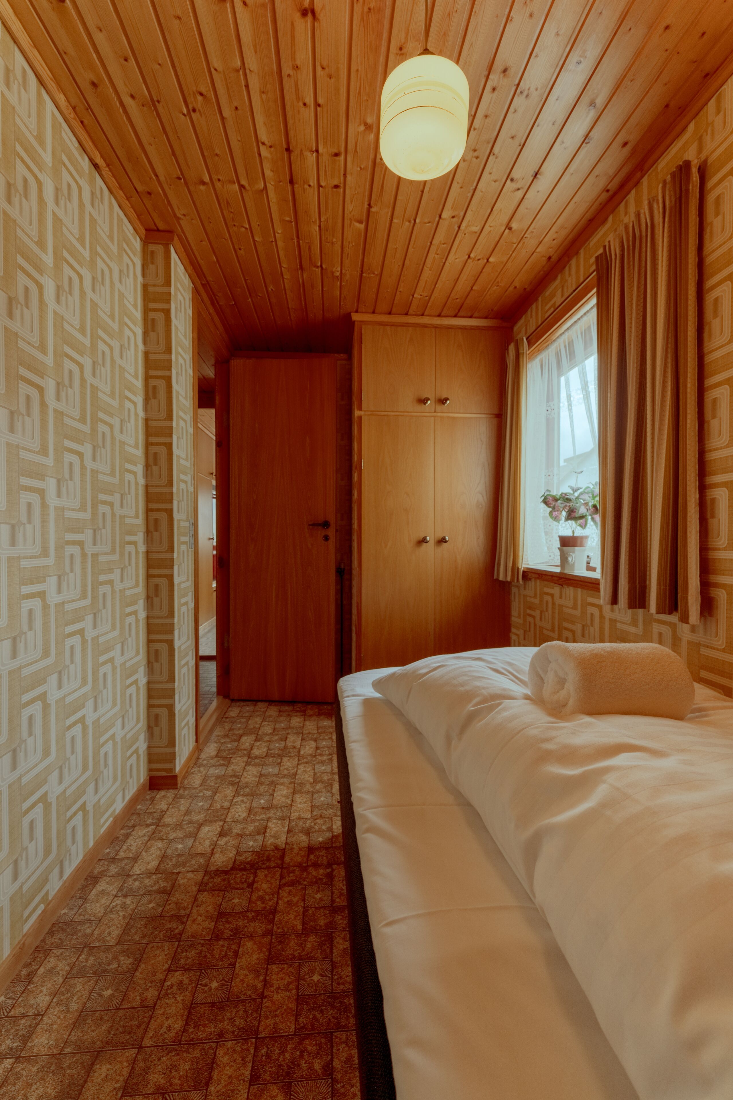 A bedroom with a wooden ceiling, a bed with white linens, a wooden wardrobe, and a patterned wallpaper.