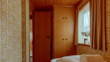A bedroom with a wooden ceiling, a bed with white linens, a wooden wardrobe, and a patterned wallpaper.