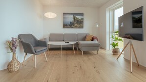 A modern living room with a grey sofa, a round coffee table, and a grey armchair. There is a large window providing natural light, a flat-screen TV mounted on the wall, and a potted plant on the floor. - New 2Br Apartment | Sea View (Tórshavn)