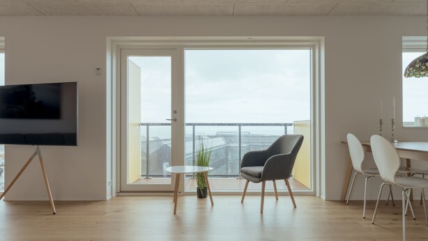 A modern living room with a flat-screen TV, a dining area with white chairs, and a balcony with a view of the city. - New 2Br Apartment | Sea View (Tórshavn)