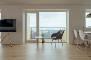 A modern living room with a flat-screen TV, a dining area with white chairs, and a balcony with a view of the city. - New 2Br Apartment | Sea View (Tórshavn)