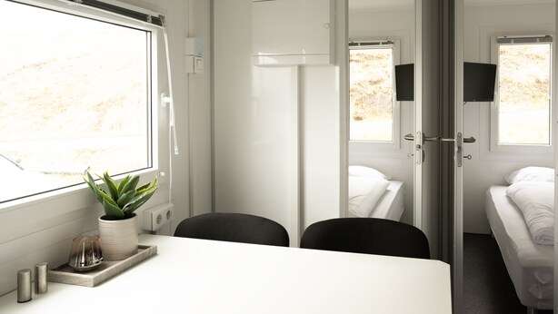A modern train compartment with a white interior, a dining table with a plant, two black chairs, a bed with white bedding, and a television mounted on the wall. - Cozy Container Rental | 3Br | 20 Min To Capital (Strendur)