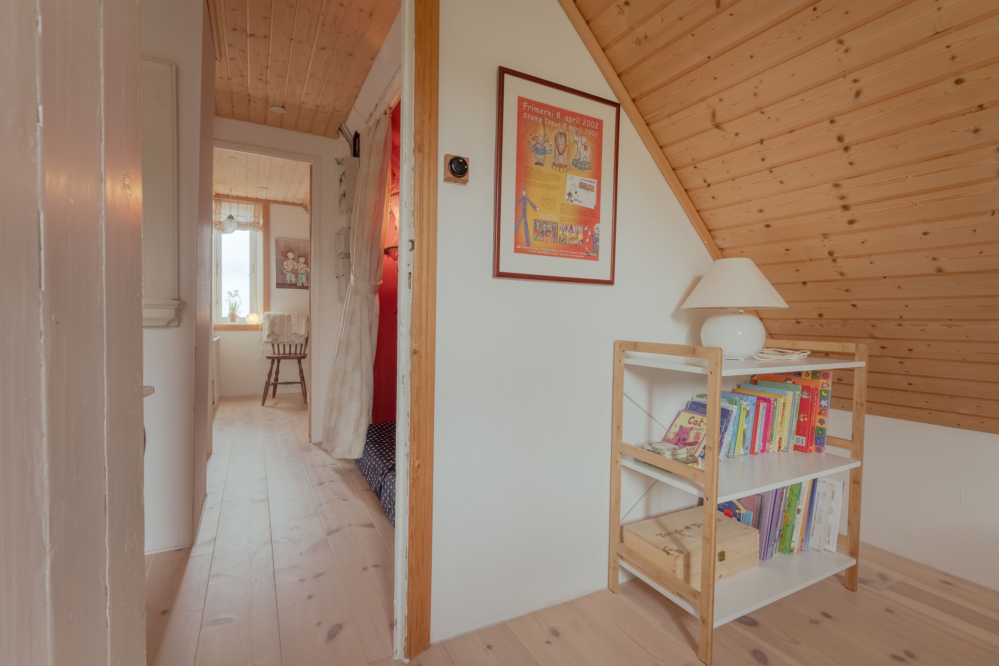 A room with a sloped ceiling, a wooden bookshelf with books, a framed poster on the wall, and a door leading to another room.