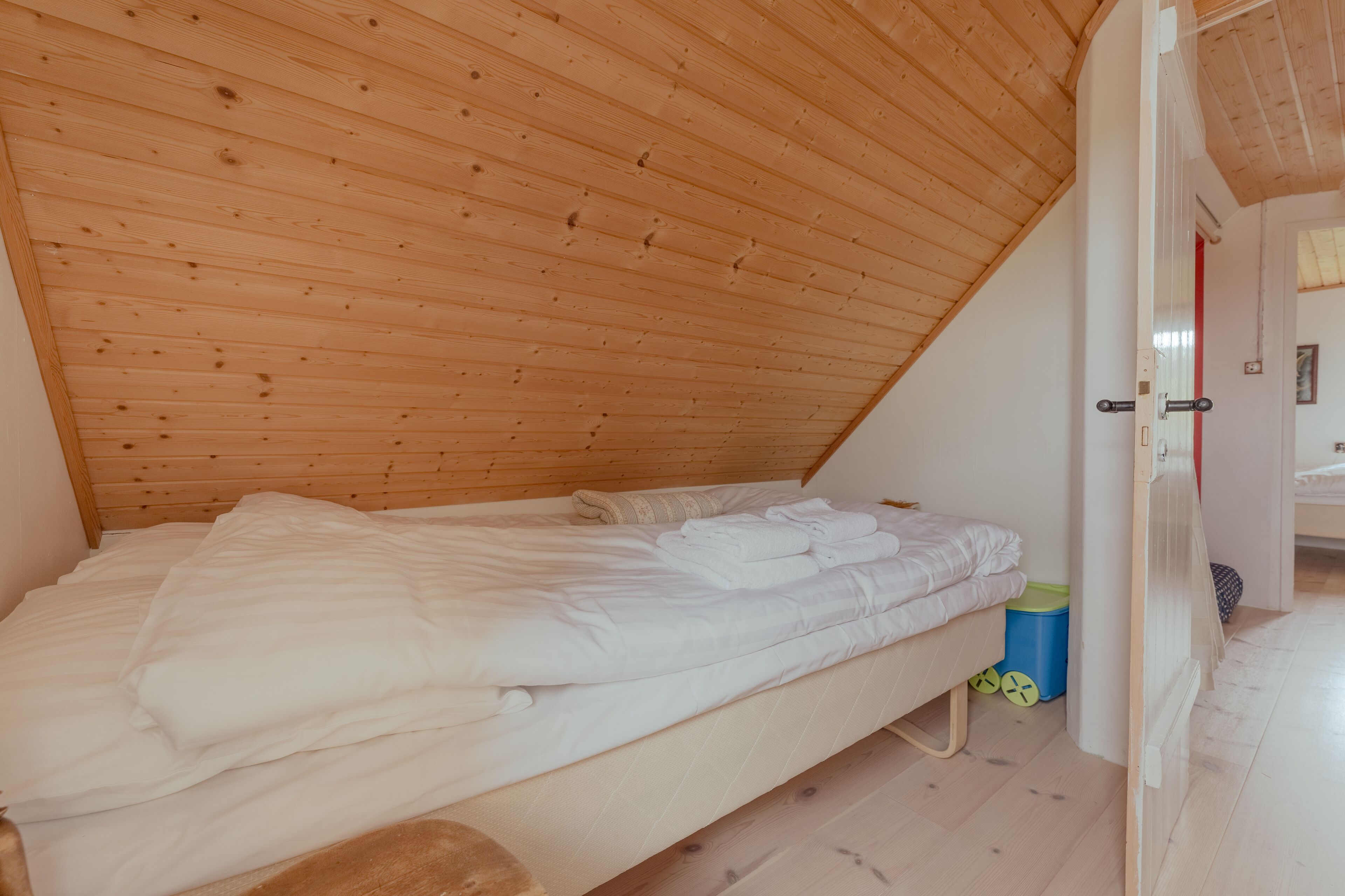 A small, single-bed room with a sloped wooden ceiling and a door leading to another room.