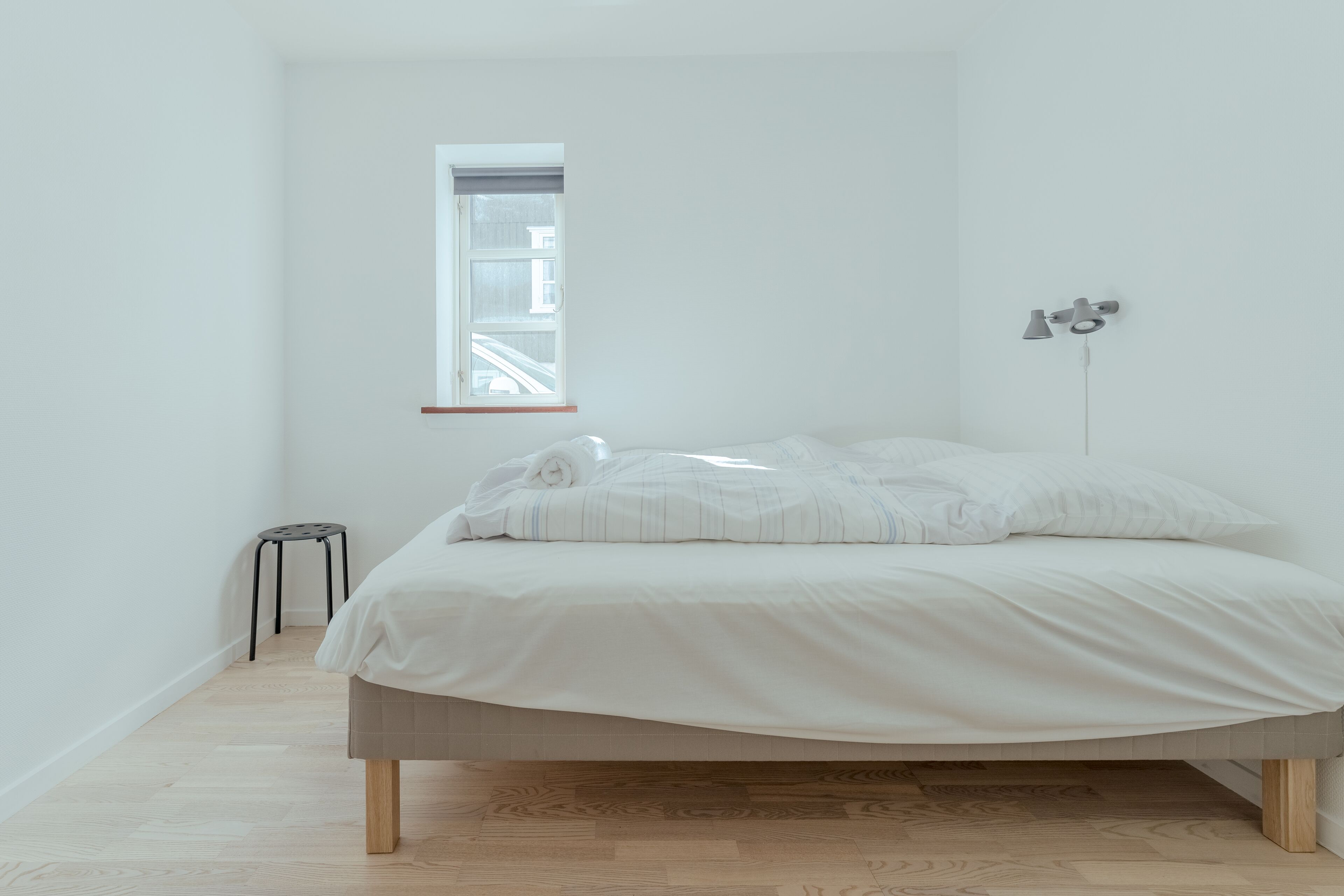 Een minimalistische slaapkamer met een bed, een klein krukje en een raam.