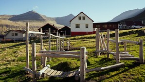 A rural setting with a white and red house, wooden play structures, and a mountain in the background. - Lovely 4-Br House | Sea View | Nature (Oyndarfjørður)