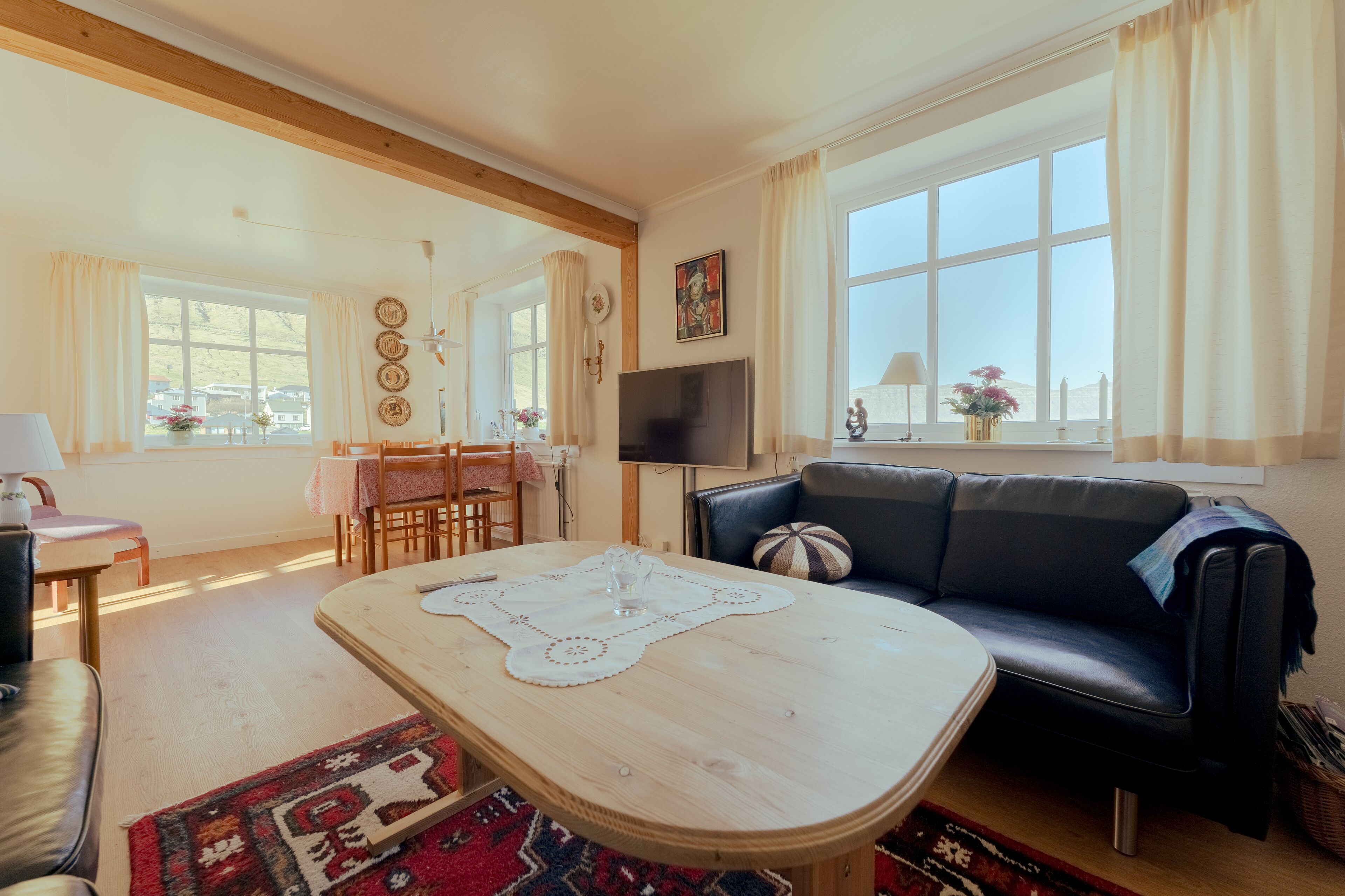 A living room with a wooden coffee table, a black leather sofa, a TV, and large windows with curtains.