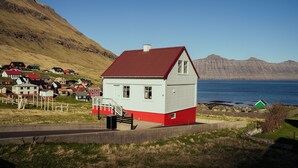 A small white building with a red roof, situated on a grassy hill overlooking a body of water and a mountainous landscape. - Lovely 4-Br House | Sea View | Nature (Oyndarfjørður)
