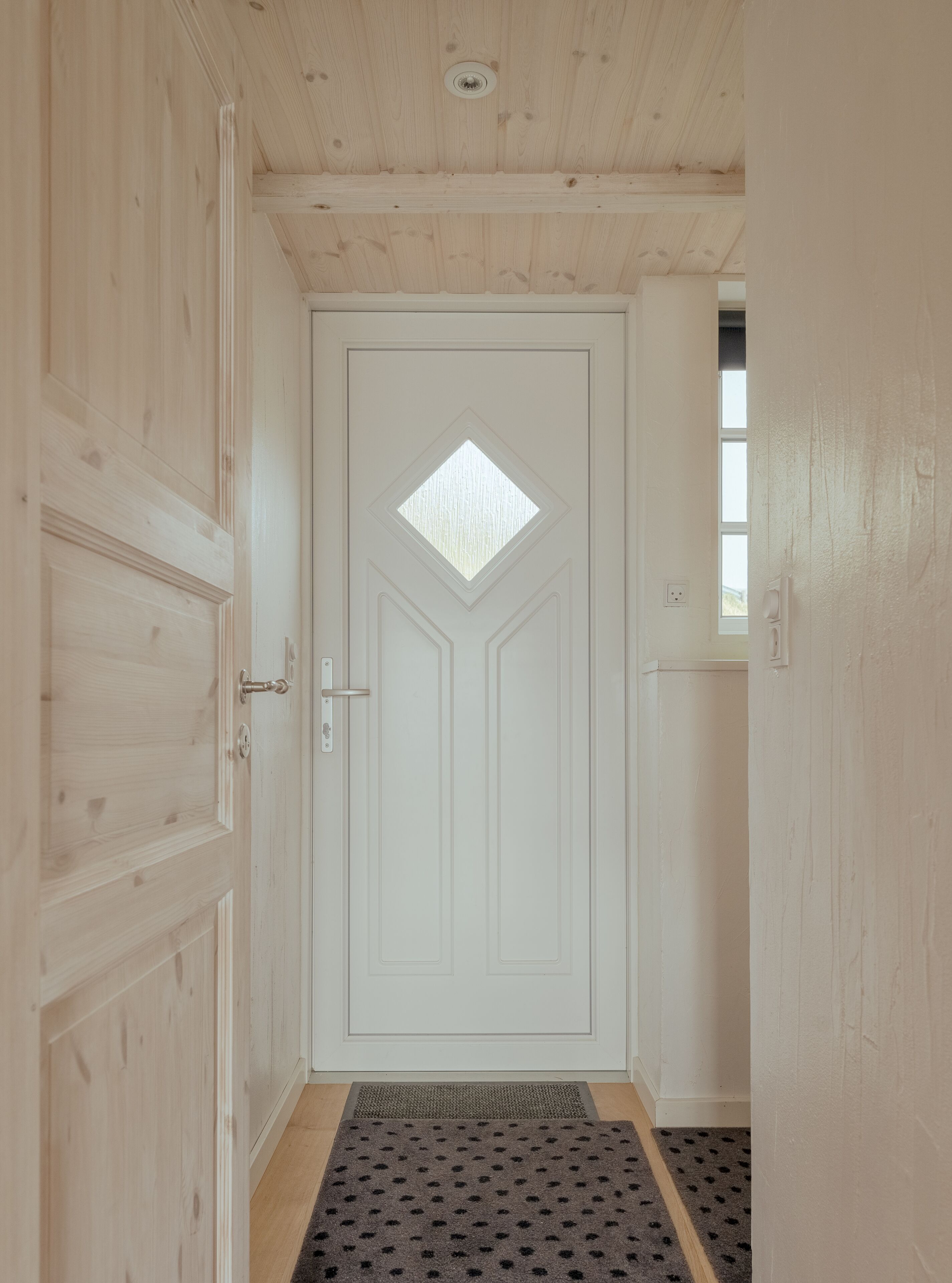 Un pasillo con una puerta blanca que tiene un ventanal en forma de diamante, un felpudo y paredes de madera.