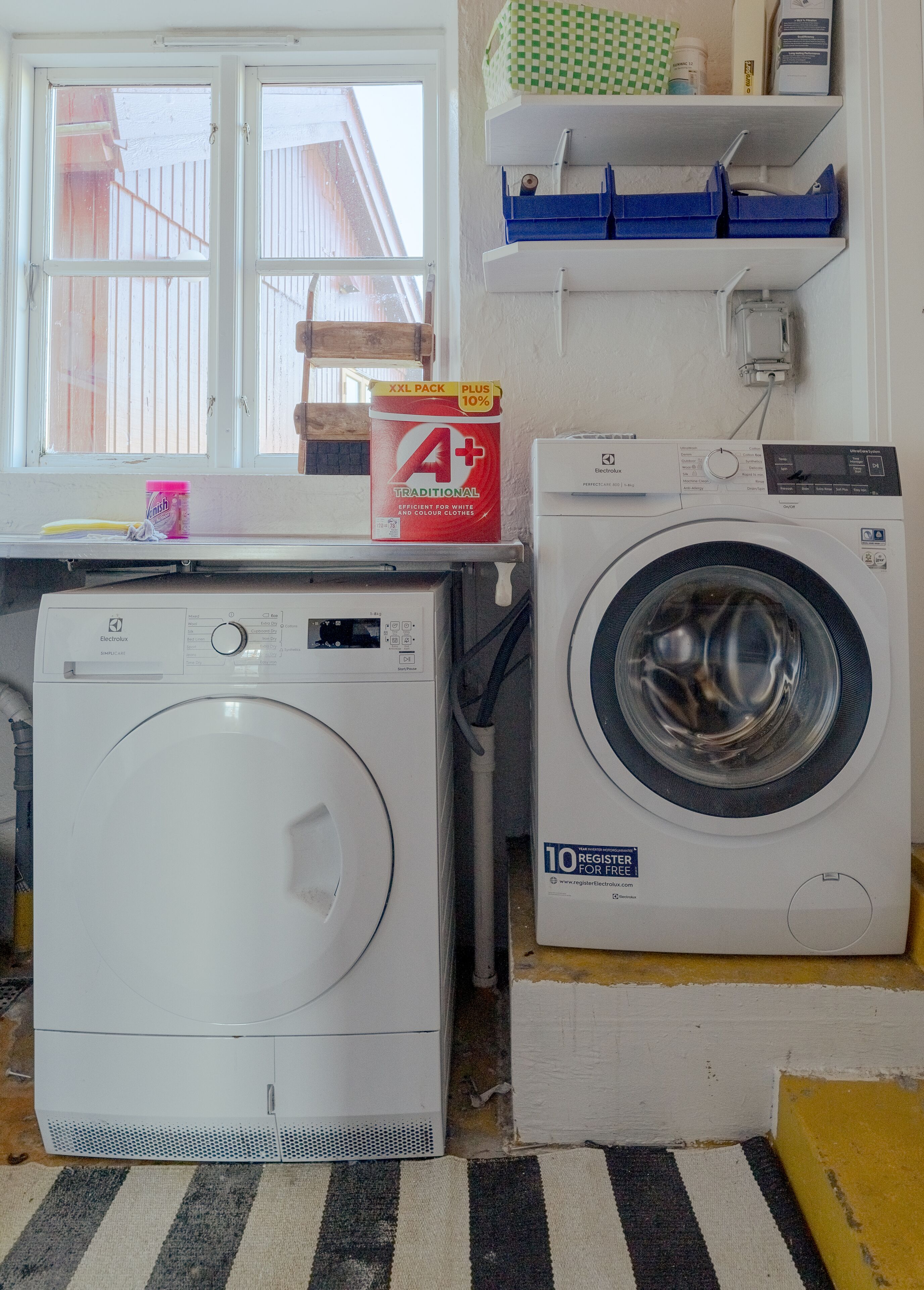 A laundry area with two washing machines, a window, and a shelf with cleaning supplies.