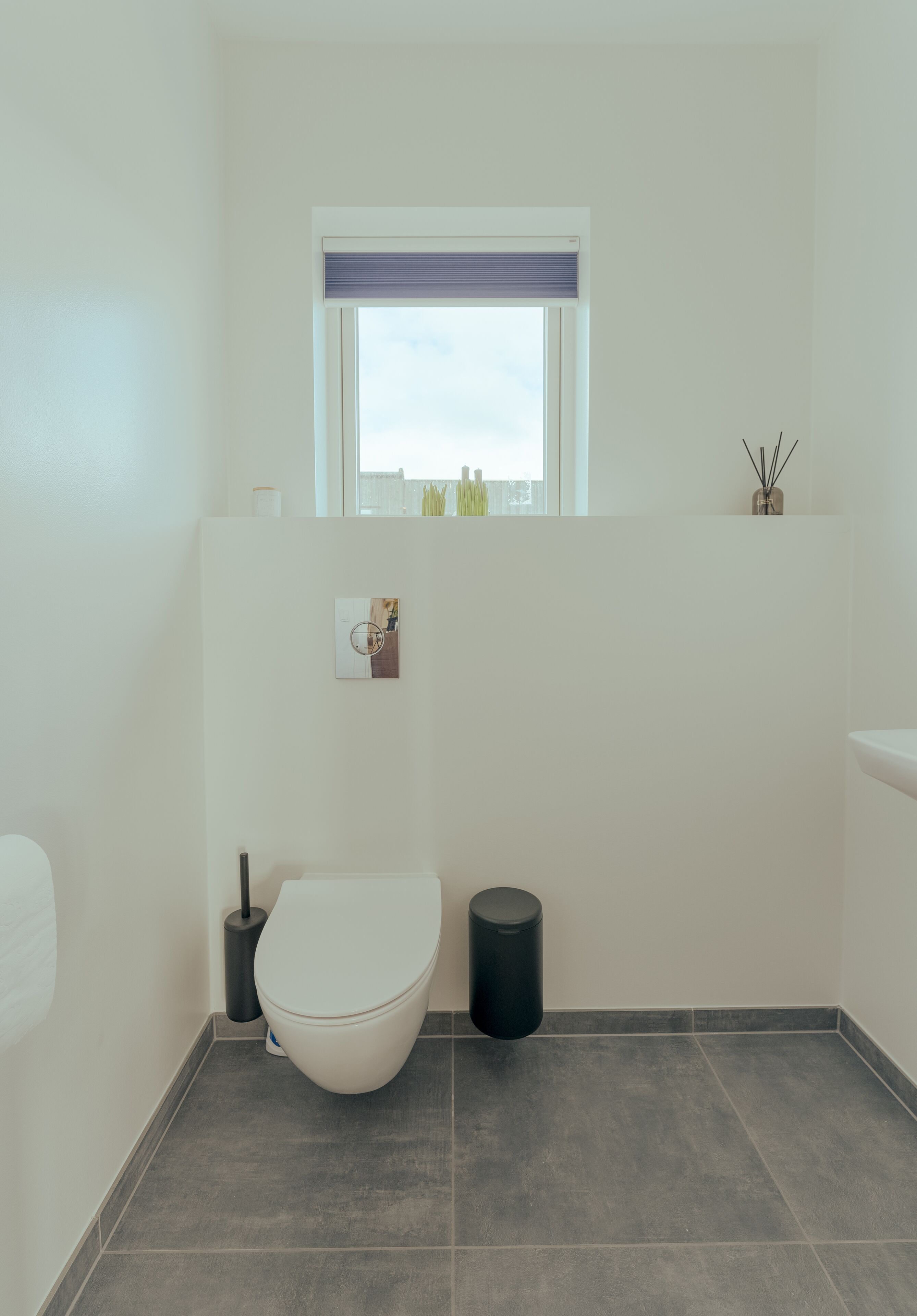 Une petite salle de bain comprenant des toilettes, une fenêtre avec un store, une poubelle et une petite étagère ornée d’objets décoratifs.