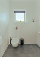 A small bathroom with a toilet, a window with a blind, a trash can, and a small shelf with decorative items.