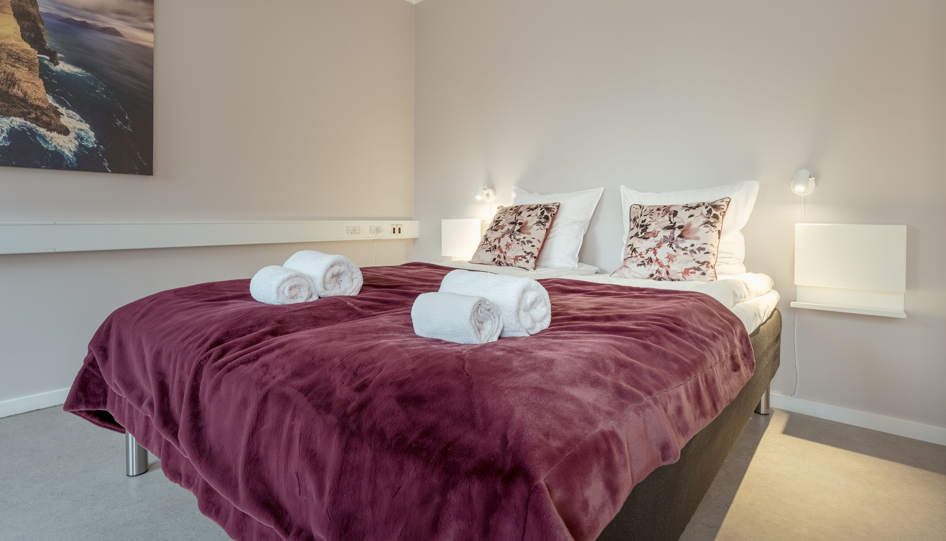 A hotel room with a large bed, two rolled towels, a red blanket, white bedding, floral pillows, and a painting of a coastal scene.