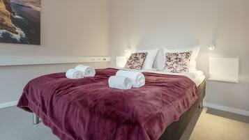 A hotel room with a large bed, two rolled towels, a red blanket, white bedding, floral pillows, and a painting of a coastal scene.