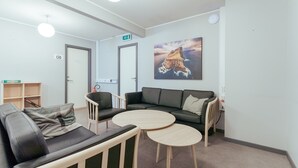 Un área de descanso moderna con un sofá, dos mesas redondas y un diván. Hay una estantería, una puerta con un letrero de salida y un cuadro en la pared.