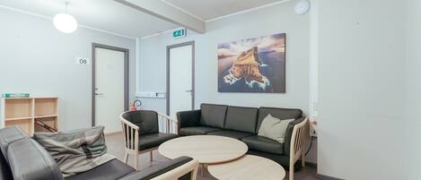 A modern lounge area with a sofa, two round tables, and a couch. There is a bookshelf, a door with an exit sign, and a painting on the wall.