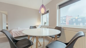 Una habitación moderna con una mesa redonda, cuatro sillas negras, una lámpara colgante morada, una ventana con vistas a edificios y un radiador.