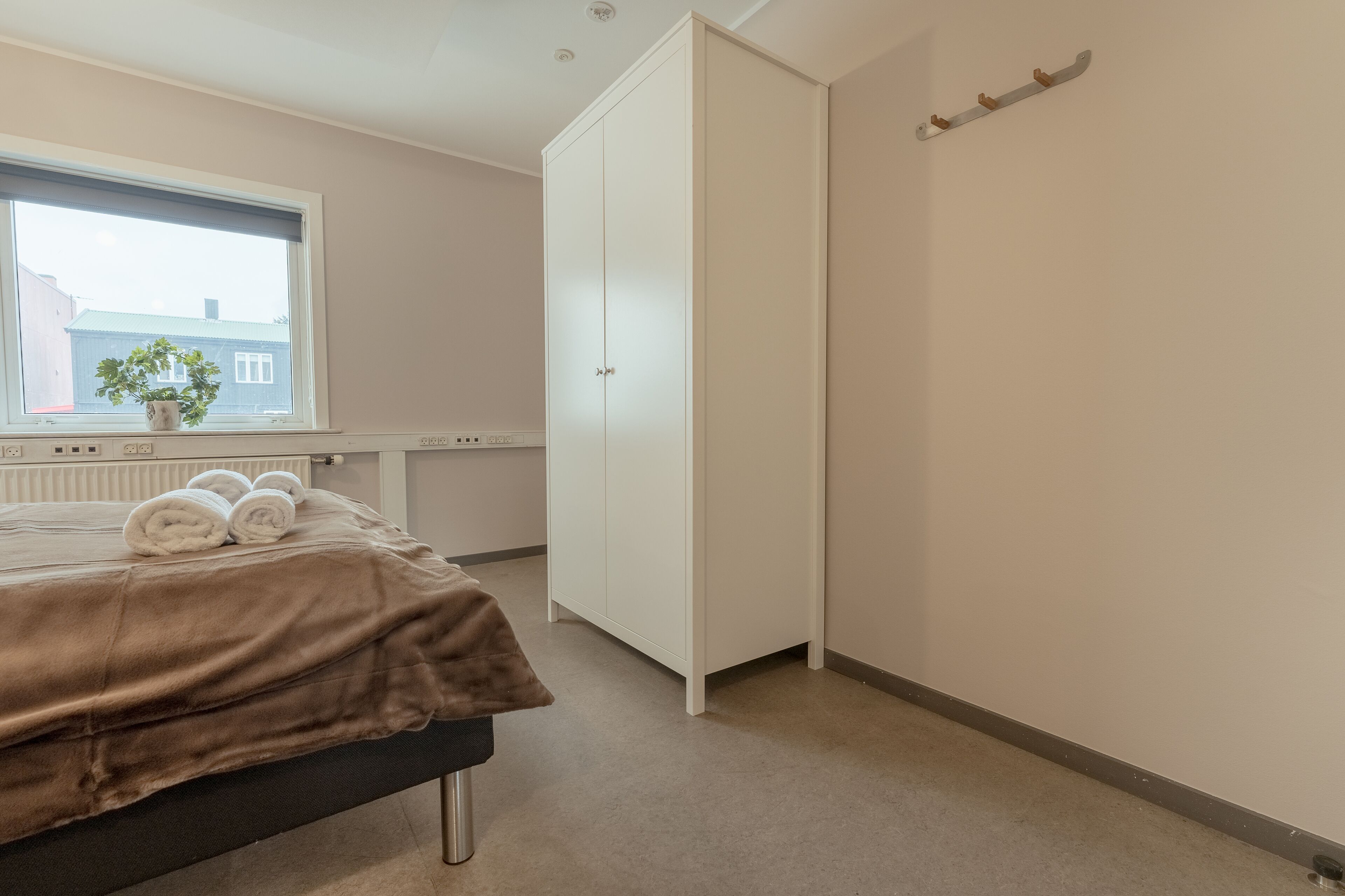 Et soveværelse med seng, radiator, et vindue med udsigt til bygninger og et klædeskab.