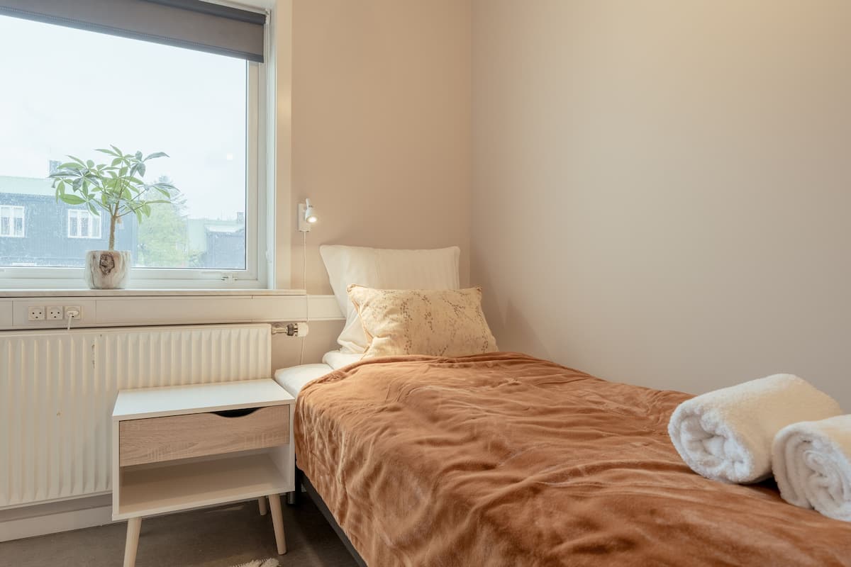 Un dormitorio con cama, mesita de noche, radiador, ventana con planta y vistas a edificios.