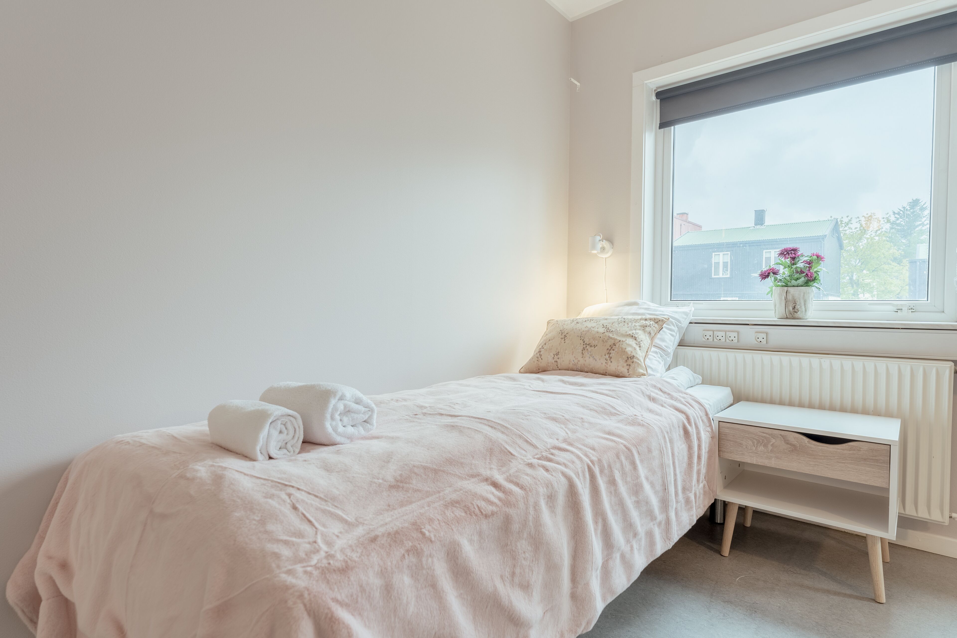 A bedroom with a bed, bedside table, and a window with a view of a house.