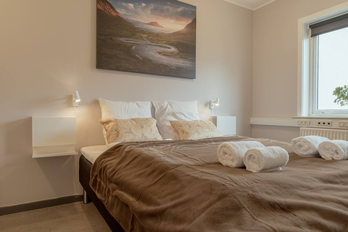 Un dormitorio con una cama grande, un cuadro de un paisaje, una ventana con vistas y una mesita de noche con lámpara.