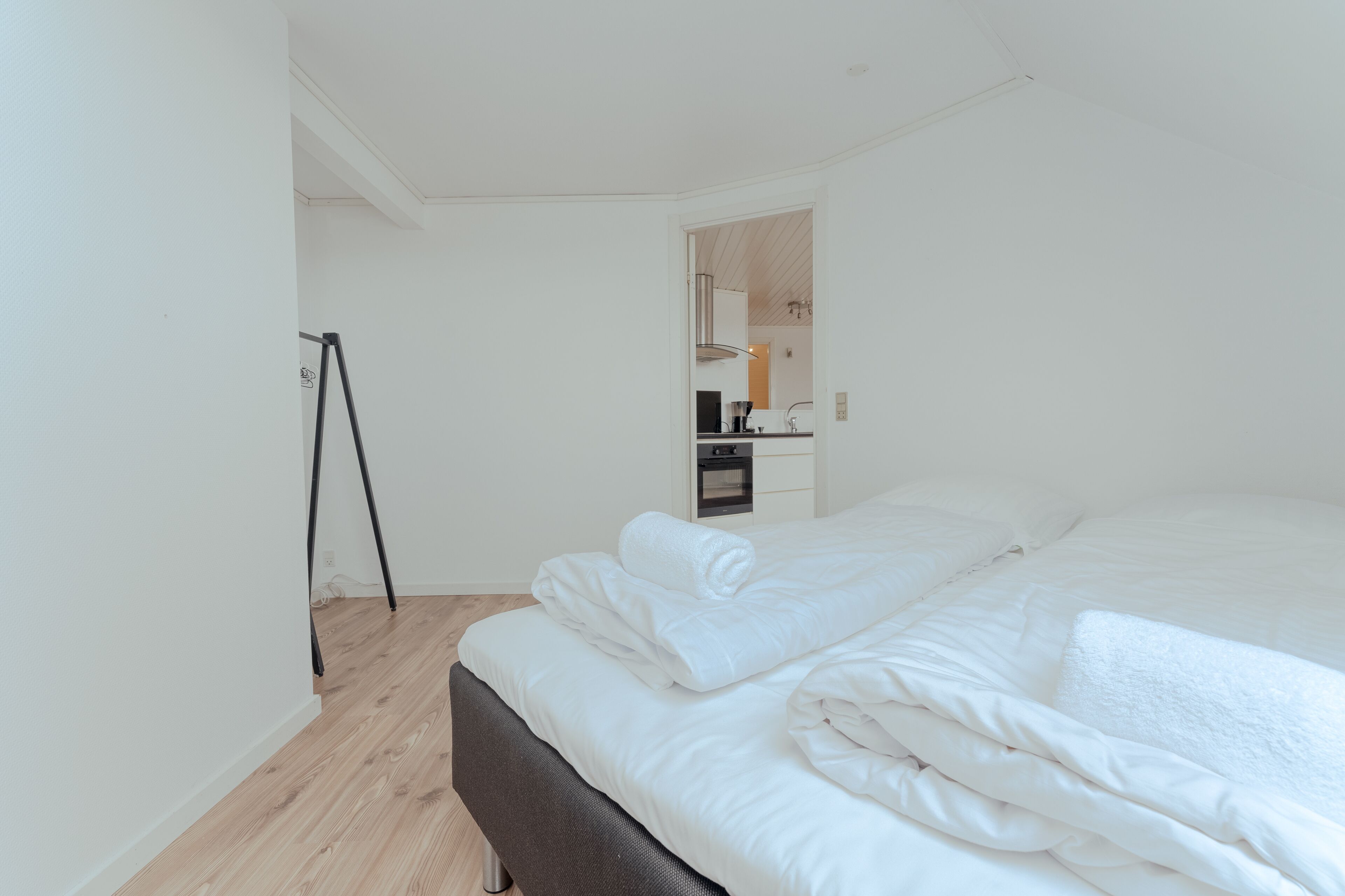 A bedroom with a neatly made bed, a wooden floor, and a view into the kitchen area.
