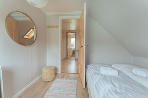 Ein Schlafzimmer mit einem Bett, einem runden Spiegel, einem gewebten Hocker und einem Flur, der in einen weiteren Raum führt.