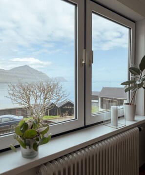 A room with a large window overlooking a body of water and mountains, a radiator, and a potted plant on the windowsill. - Serenity By The Sea | 3-Br House | Sea View (Sydrugota)