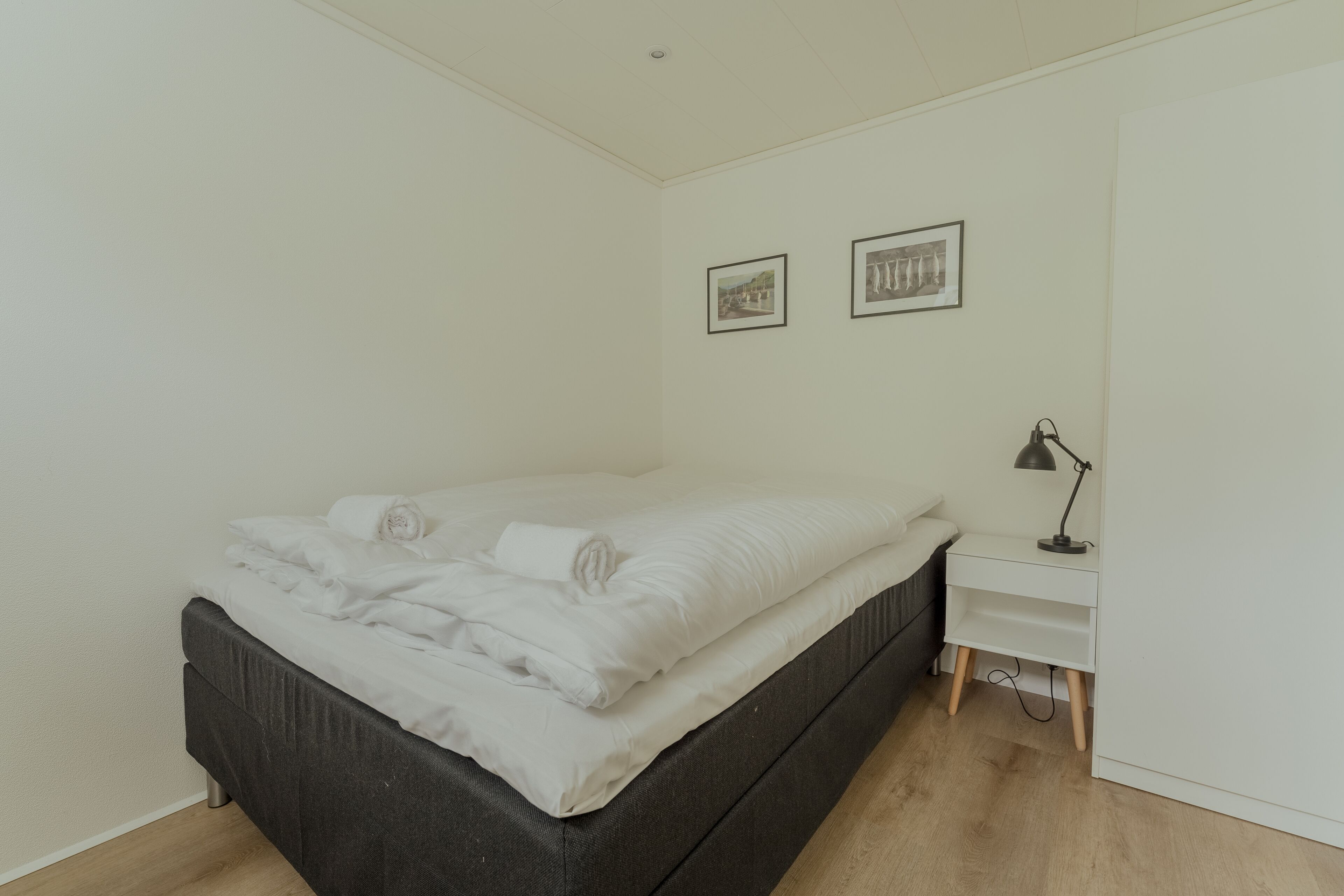 A bedroom with a bed, a bedside table with a lamp, and two framed pictures on the wall.