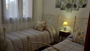 A bedroom with two beds, a bedside table with a lamp, and a window with sheer curtains.
