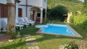 A pool with a stone edge, surrounded by a grassy area with stepping stones, white lounge chairs, and a stone pillar.