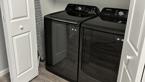 Une buanderie avec deux machines à laver et deux sèche-linge noirs, une porte blanche et une étagère en métal au-dessus.