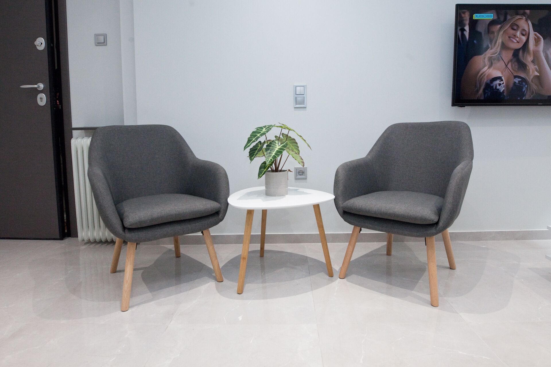 Two grey armchairs with wooden legs, a white table with a potted plant, and a TV on the wall.