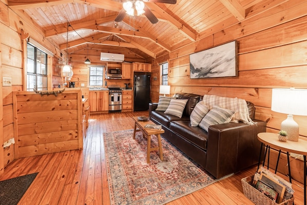 Un acogedor interior de cabaña con paredes de madera, un sofá, una mesa de centro, una zona de cocina y una mesa de comedor.