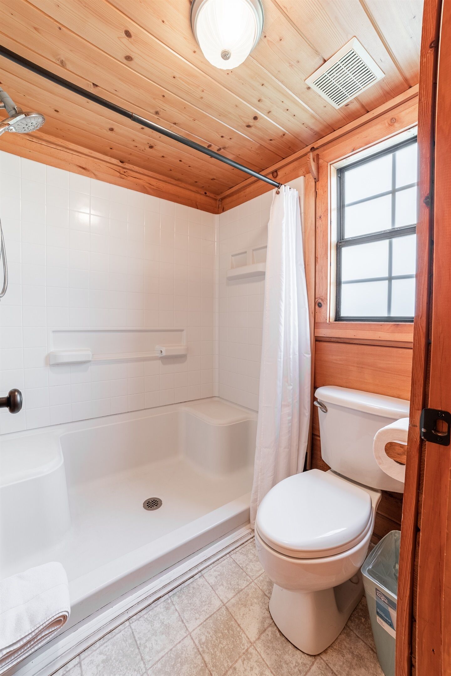 Un baño con ducha, inodoro y lavamanos, con paredes de madera y una ventana.