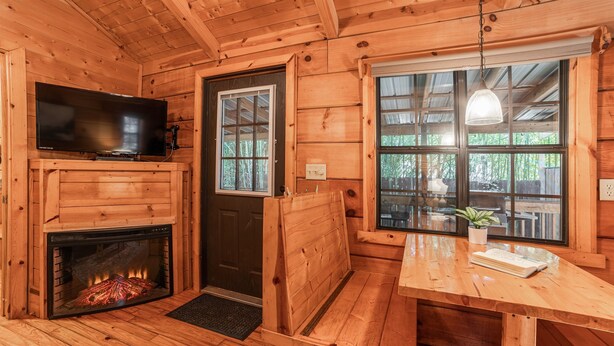 A wooden cabin interior with a fireplace, a television, a bench, and a table with a plant. - Cozy Cabin w/ Hot Tub & Fenced in Yard for Pets (Hot Springs)