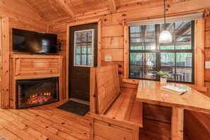 A wooden cabin interior with a fireplace, a television, a bench, and a table with a plant. - Cozy Cabin w/ Hot Tub & Fenced in Yard for Pets (Hot Springs)