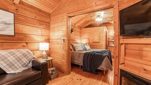A cozy wooden cabin interior with a bed, a leather sofa, a television, and a fireplace. - Cozy Cabin w/ Hot Tub & Fenced in Yard for Pets (Hot Springs)