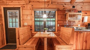 A wooden cabin interior with a central wooden bench table, a window with a view, and a kitchen area with a sink and dish rack. - Cozy Cabin w/ Hot Tub & Fenced in Yard for Pets (Hot Springs)